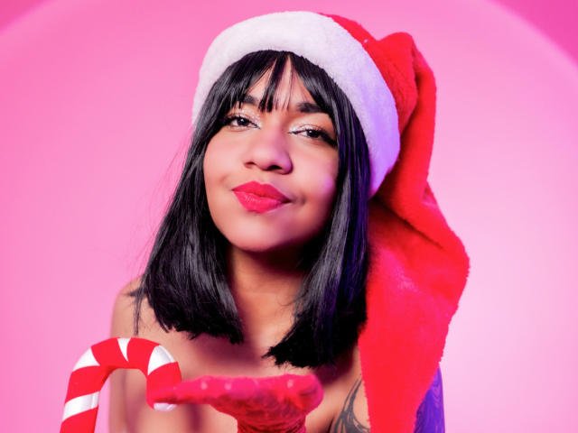 MaddyMagic with black hair, red Santa hat, holding candy cane, pink lipstick, slight smile, tattoo visible on right shoulder, pink background, cute, sexy, festive, soft lighting.
