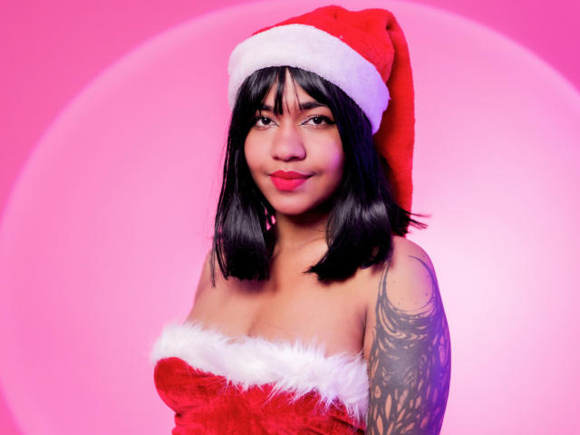 MaddyMagic with black shoulder-length hair, wearing a red Santa hat and strapless red fur-lined top, showing tattoos on her right arm, and a slight smile. Pink background.