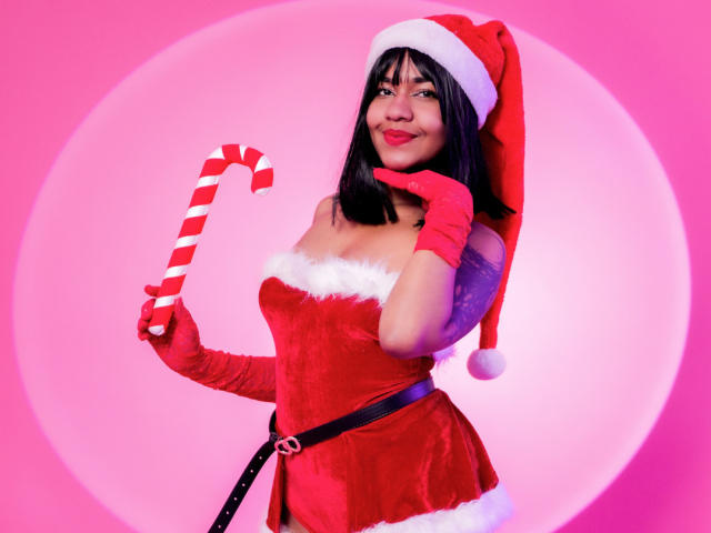 MaddyMagic in a red Santa dress, holding a candy cane, wearing a Santa hat and red gloves, with her hand near her mouth, smiling. Her tits are visible.