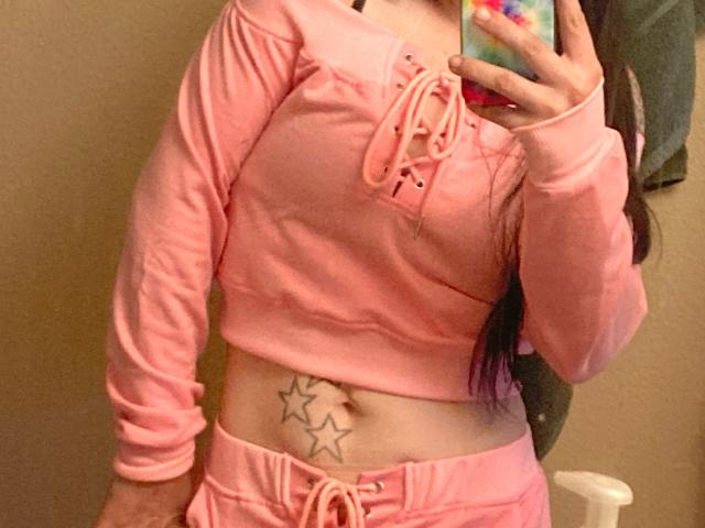 Nikkiy69 in pink long-sleeve crop top and matching pants, showing a tattoo of three stars on her lower belly, taking a mirror selfie with a colorful phone case.