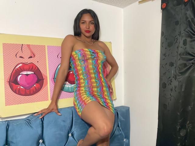 BellaChaudAmour, with dark hair and red lipstick, wears a rainbow-striped, strapless, tight dress, showing her ass, standing on a blue couch, hands on her hips.