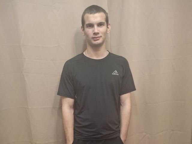 Noctyve with short brown hair, wearing a black Adidas t-shirt, standing against a beige curtain, hands in black pants, smiling slightly, looking at camera.