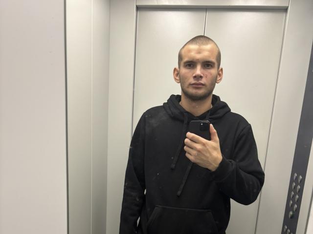 Noctyve, bald, black hoodie, holding phone, standing in elevator, looking at camera, slight frown, right hand on phone, left hand in pocket, neutral expression, short beard.