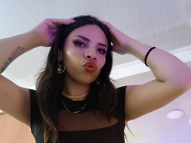 VioletaStone a des paupières violettes gonflées, des lèvres rouges brillantes et des cheveux noirs ondulés, elle porte un haut noir transparent, un collier en or et des boucles d'oreilles, avec les mains dans les cheveux, et regarde l'appareil photo..