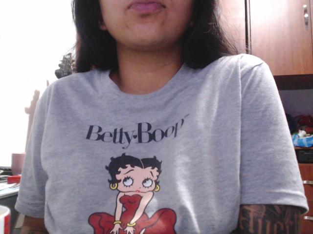 Unicornia69 wearing a gray t-shirt with a Betty Boop graphic, showing her tits, with black hair, pink lipstick, and a tattooed arm, in a kitchen.