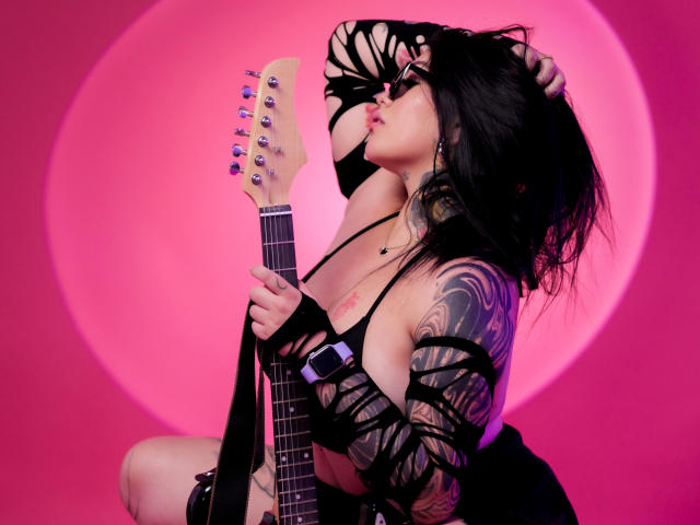 TessaMagic, con cabello negro, tatuajes y un top negro con aberturas, sosteniendo una guitarra, con los senos expuestos, en una toma con luz rosa, con un dedo en la boca, luciendo seductora..