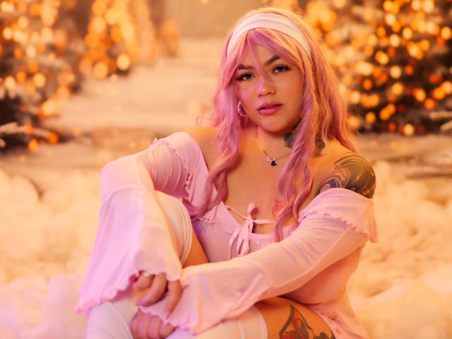 TessaMagic, com cabelo rosa, faixa branca na cabeça e blusa rosa com um ombro descoberto, sentada com as pernas cruzadas, mostrando tatuagens no braço direito e na coxa esquerda, em uma iluminação suave e quente..