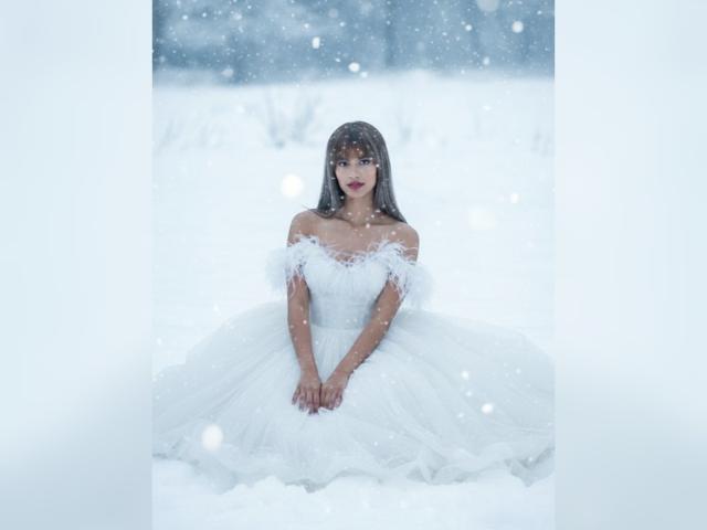 IsabelaLoben in a white off-shoulder wedding dress with puffy sleeves, sitting in snow, hands together, snowflakes falling, dark long hair, red lipstick, looking forward.