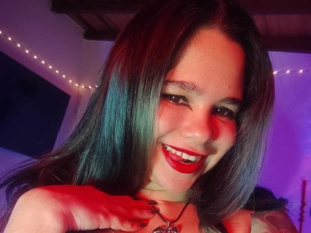AshGraven has dark green-blue hair, red lipstick, and a playful smile with red and purple lighting. She's touching her chest with black-painted nails. Her tits are slightly visible.