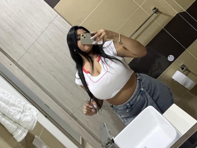 AbrilLuxurie met lang zwart haar, die een witte crop top draagt die haar grote borsten en een roze bh laat zien, en hoge jeans, die een spiegelzelfportret maakt in een badkamer..
