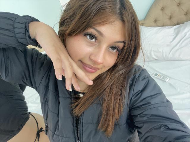 BrendaGreen, with brown hair and black puffy jacket, sits on bed, showing a slight smile, touching her chin, black underwear visible, remote control on white pillow.