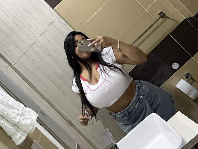 Abril, met lang zwart haar, grote borsten, een witte crop top, een rode bh, blauwe jeans, die een telefoon vasthoudt, een spiegelzelfportret, in een badkamer, met bruine tegels, een witte handdoek, een armband..