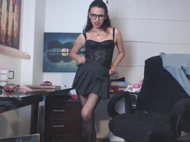 AfroditaRgenis in a black lace corset, pleated skirt, fishnet stockings, and glasses, standing with red lipstick, pulling up her skirt, showing her tits, in a messy room.