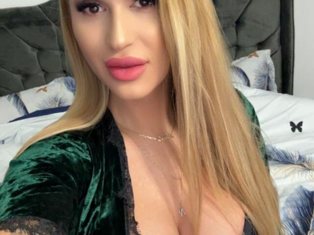 NicolSNaya with blonde hair, green velvet robe, black lace trim, pink lips, necklace, and visible large tits. She's looking at the camera with a slight smile.