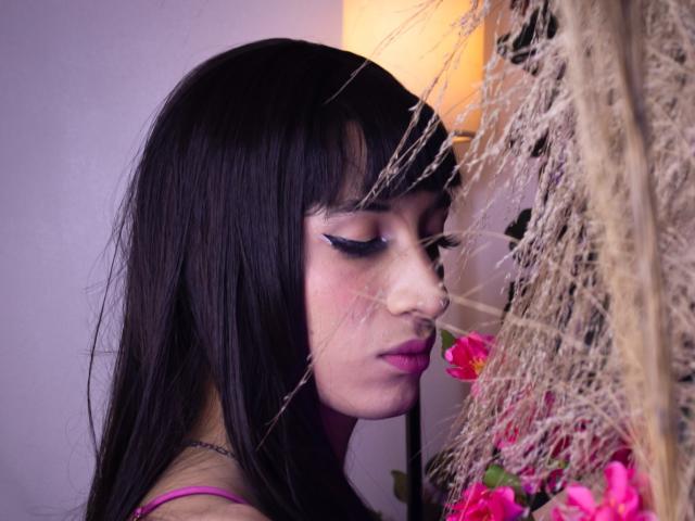 Close-up of AlexiaHearts with straight black hair and bold pink lipstick. She has dramatic black eyeliner with winged tips. Her eyes are closed, and she's leaning against a wall with dried grass and pink flowers. Soft purple and yellow light in the background. Her face is slightly shadowed, with a thin necklace visible. The composition focuses on her profile, with the floral elements and grass adding texture.