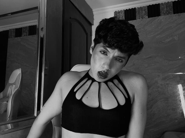 EllisBrown, avec des cheveux courts et bouclés, les lèvres pincées, portant un soutien-gorge de sport noir avec découpes au niveau de la poitrine, assise sur une chaise, regardant la caméra, sur une photo en noir et blanc..