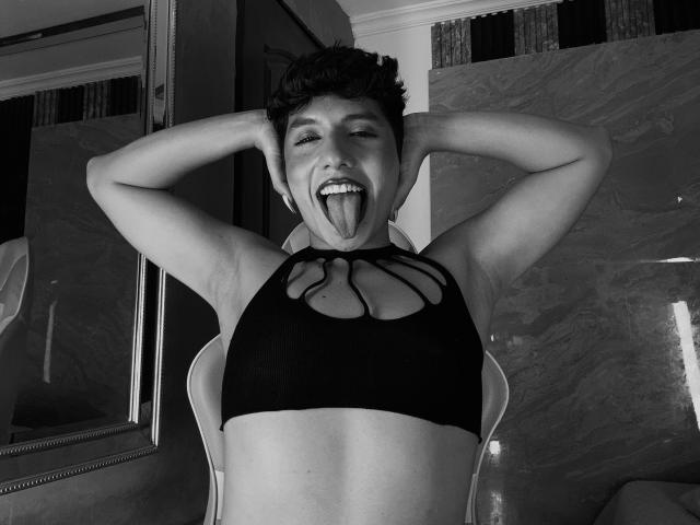 EllisBrown, avec ses courts cheveux foncés, les bras levés, la langue tirée, portant un soutien-gorge de sport noir découpé, montrant de petites seins, assise sur une chaise, photo en noir et blanc..