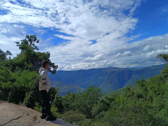 DareckD stands on a rocky cliff, facing mountains with a blue sky and white clouds. Wearing a white hoodie and black pants, DareckD looks over the green landscape.