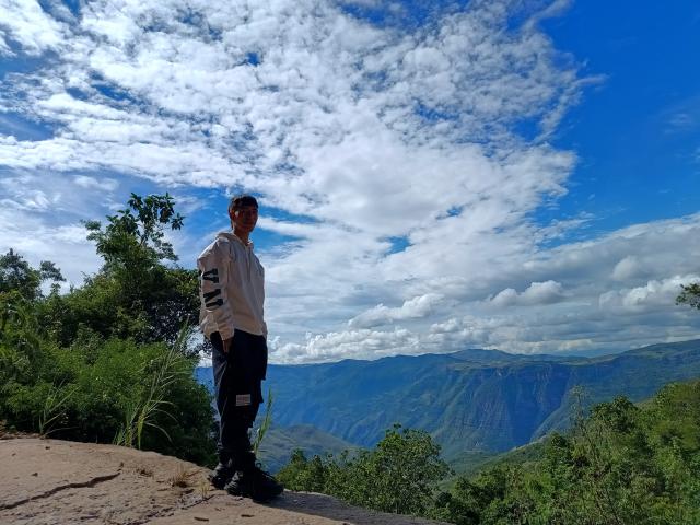 DareckD stands on a rocky cliff, wearing a white hoodie with black stripes and black pants, looking at the blue sky with scattered white clouds.