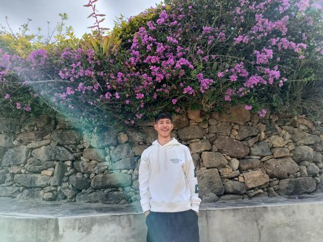 DareckD stands in front of a stone wall, wearing a white hoodie and black pants, with purple flowers above. He's smiling with hands in pockets, looking cute.