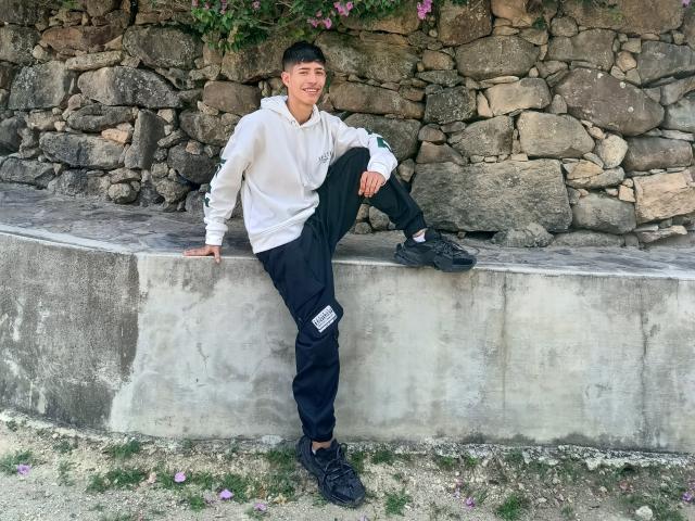 DareckD, sitting on a concrete ledge, wearing a white hoodie, black pants, and black sneakers, with one leg up, smiling, against a stone wall.
