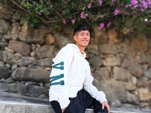 DareckD with short black hair, smiling, wearing a white hoodie with green stripes and black pants, sitting on a stone bench, purple flowers in the background.