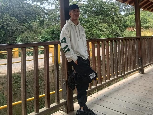 DareckD leans against a wooden railing, wearing a white hoodie, black pants, and a green cap, with a black bag in hand, smiling at the camera.