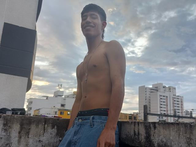 Shirtless DareckD with a slight tan, black cap, necklace, and blue jeans stands on a rooftop, smiling, with his hands by his sides. Sky and buildings in background.