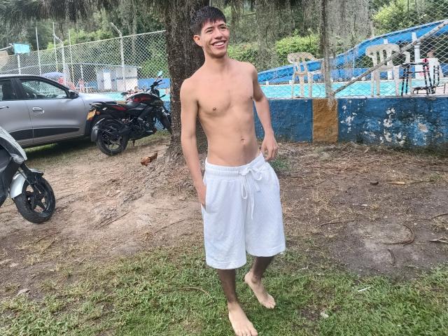 DareckD is shirtless, smiling, with white shorts, hands in pockets, barefoot, standing on grass, next to a tree, with a motorcycle and car in the background.