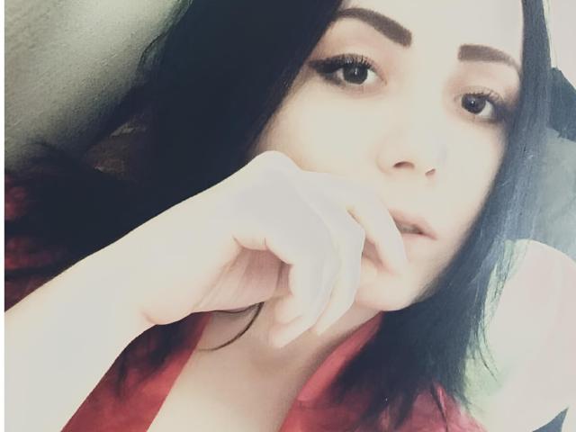 NyxKiss with black hair, thick eyebrows, long eyelashes, and red shirt, touching her slightly open mouth with a finger. Her expression is relaxed and slightly seductive.