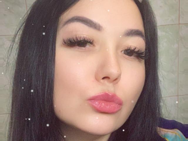 NyxKiss has black hair, thick eyebrows, long eyelashes, and pink glossy lips pouting. Snow-like dots are on the image, and she has a small nose ring.