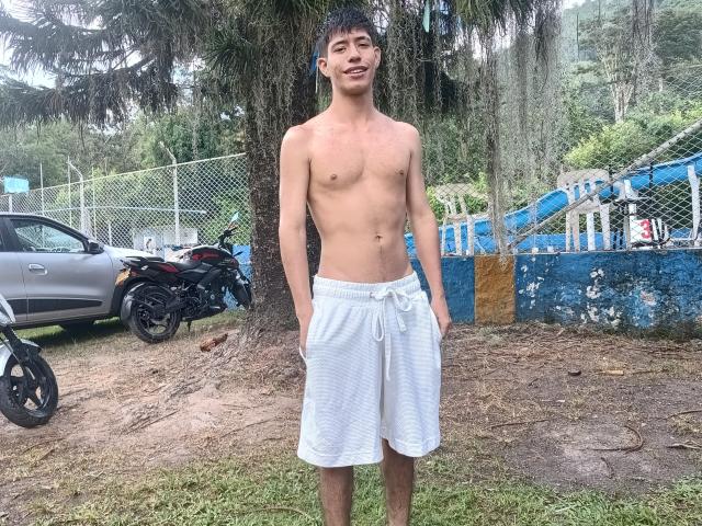 Shirtless DareckD with white shorts, standing, hands in pockets, slight smile, short dark hair, slim build, small nipples, small belly button, outdoors, near a tree and motorcycle.