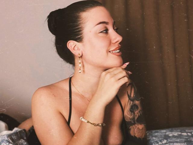 MiaKhallirae with her dark hair in a bun, smiling, holding her chin, showing off her tattooed arm, wearing a black halter top, and gold earrings.