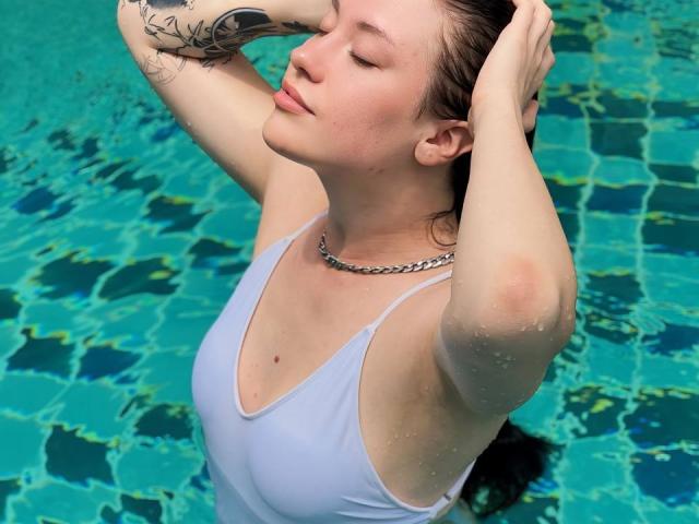MiaKhallirae with wet white tank top, arms raised, hands in brown hair, blue-green water background, visible tattoo on right arm, chain necklace, eyes closed, lips slightly parted.
