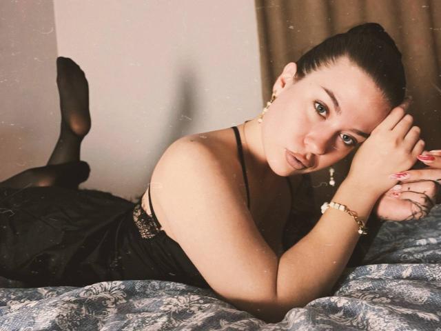 MiaKhallirae laying on a bed, wearing black lingerie and sheer stockings, with her ass up and her tits down, looking at the camera with a serious expression.