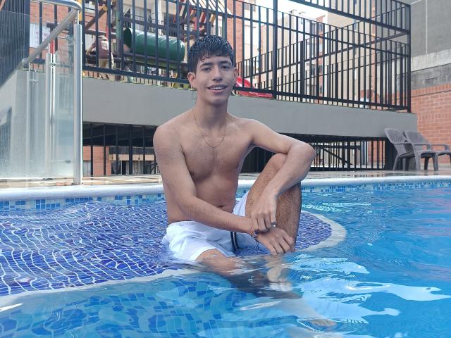 DareckD, shirtless, with wet brown hair, white swim trunks, sitting in a blue-tiled pool, smiling, hands on knee, water up to thighs, black metal railing in background.