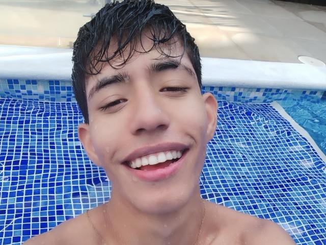 DareckD's wet, short black hair, smiling face with visible teeth, and small nose are in a blue-tiled pool, wearing a gold necklace, looking happy.