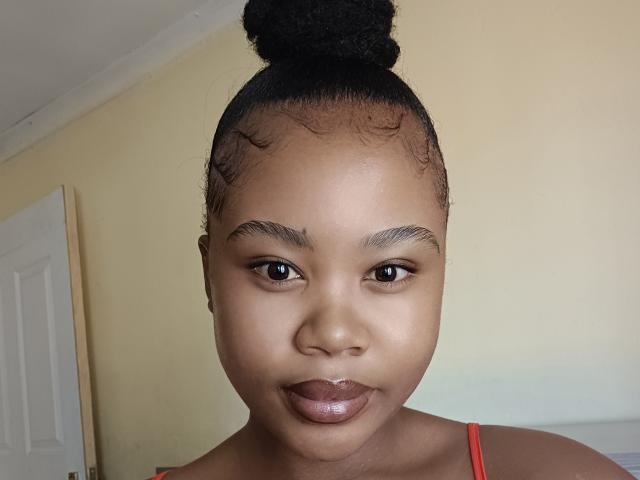 Brielly has dark brown skin, a top bun hairstyle, and small eyebrows. She has full, shiny lips and is wearing a red strap. Her eyes are dark and she's looking at the camera.