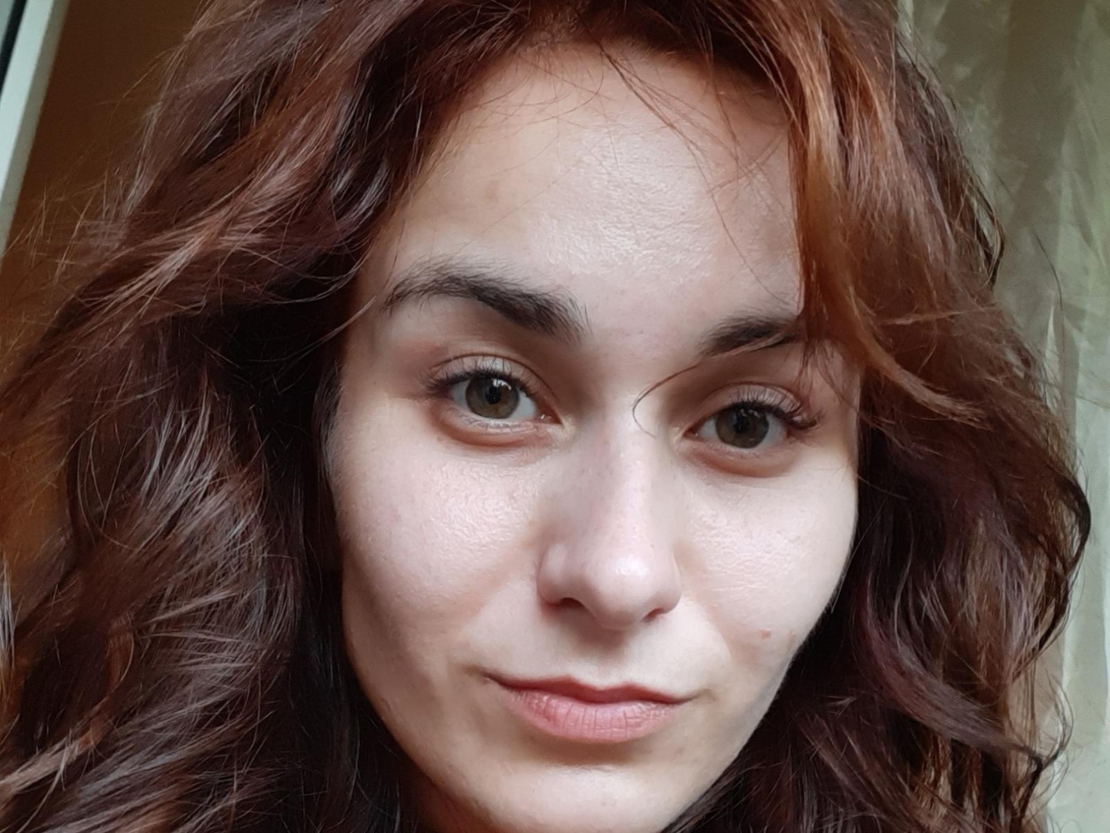 SnuggleBelly has wavy, reddish-brown hair, fair skin, and brown eyes with a subtle smile. She's looking directly at the camera with a slight, confident expression.