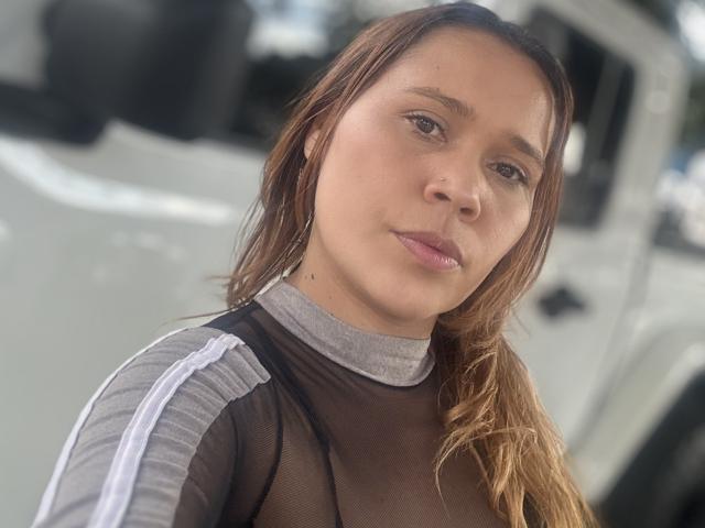 Close-up of SophiaQueens with light brown skin, brown wavy hair, and green eyes. She has subtle makeup with pinkish lips. She wears a black mesh top with gray and white ribbed sleeves. The background is blurred, showing a white vehicle. The image has natural light, highlighting her facial features and top textures. Her expression is neutral, slightly curious. The composition centers on her face, with a slight right angle.