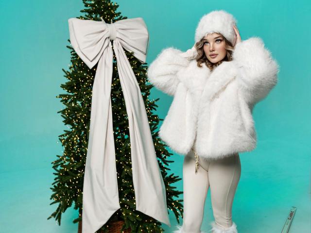 FabulousMagic with white fluffy coat, hat, and white pants, standing next to a Christmas tree with a large white bow, blue background. Her ass is covered by the coat.