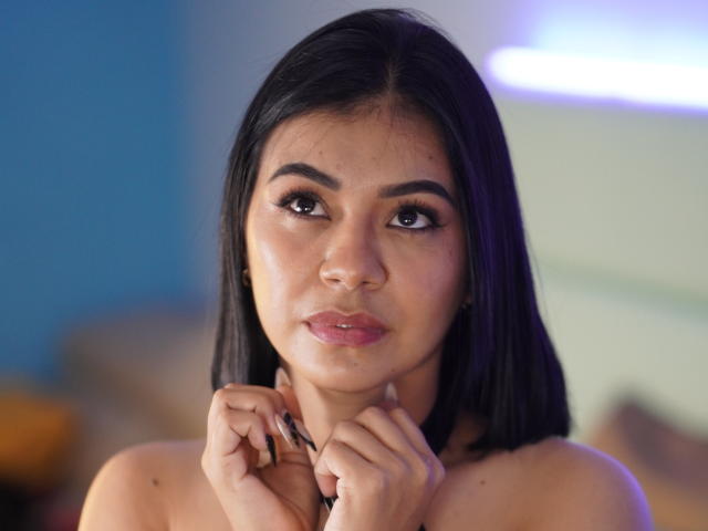 AngelVega has medium brown skin, black hair, and full pink lips. She's looking up with her hands near her neck, showing her tits and dark, glossy eyes.