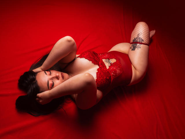 SwettieSammy con i suoi seni in pizzo rosso, braccia sopra la testa, occhi chiusi, sdraiata su un tessuto rosso, calze a rete sulla gamba destra, tatuaggio sulla coscia sinistra, capelli neri..