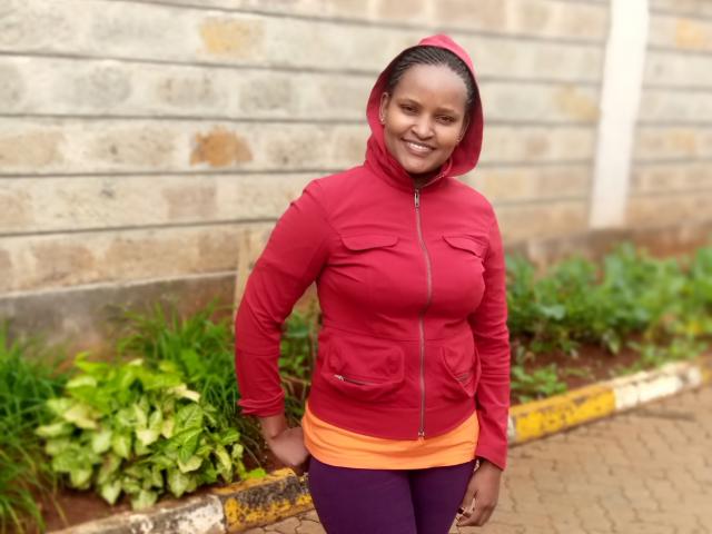Emphi69 stands in front of a brick wall, smiling. She wears a red hooded jacket with orange and purple pants. Her dark hair is pulled back, and she has small earrings. Green plants and a yellow curb are in the background. The texture of the brick wall is rough, and the curb is weathered. The image is a photograph.