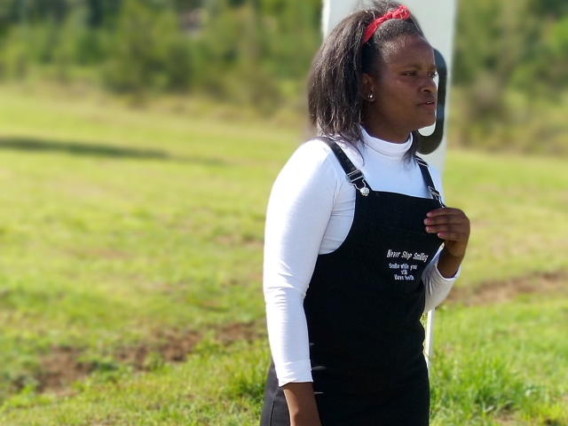 Emphi69 stands in a grassy field, facing slightly to the right. She has dark skin and wears a black apron over a white long-sleeve shirt. Her hair is tied back with a red headband. Her right hand rests on her chest. The apron has white text on it. The background is blurred, with green grass and a white pole. The light is bright, highlighting her figure. She looks focused and determined. The image is clear and well-composed.