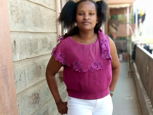 Emphi69 stands in a hallway, wearing a purple sleeveless top with lacey, floral patterns on the shoulders and white pants. Her black, curly hair is styled in pigtails. She has a neutral expression and her hands rest on her hips. A colorful bracelet is on her right wrist. The concrete wall on the left is textured and aged. The right side has a metal railing. The image is a photograph.