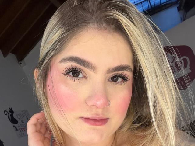 AlannaFoxy has blonde hair, pink blush on her cheeks, a nose ring, and long eyelashes. She's looking at the camera with a slight smile, her hand near her head.