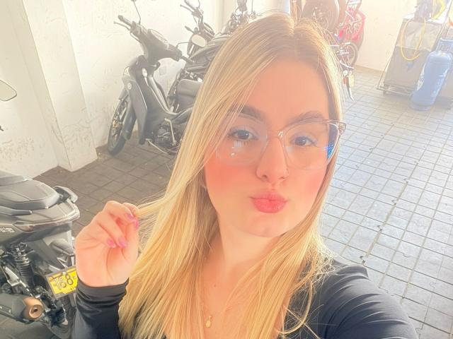 Blonde AlannaFoxy with puffy red lips, glasses, and pink nails, puckering for a kiss, holding her hair. Black top, motorcycles, and yellow sign in background.