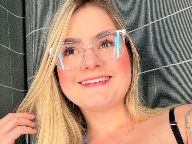 AlannaFoxy with blonde hair, clear glasses, pink blush, smiling, holding hair, black strap, visible tattoo on right shoulder, pink lipstick, white teeth.