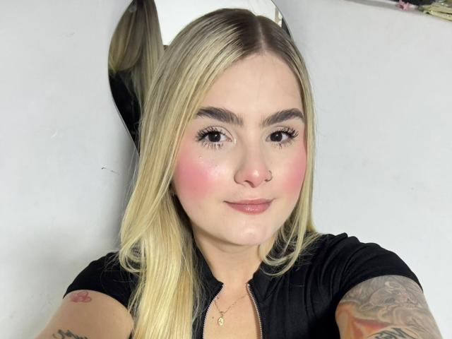 AlannaFoxy has blonde hair, pink blush, black shirt, tattoo on right arm, and a nose ring, smiling with eyeliner and long lashes, wearing a gold necklace.